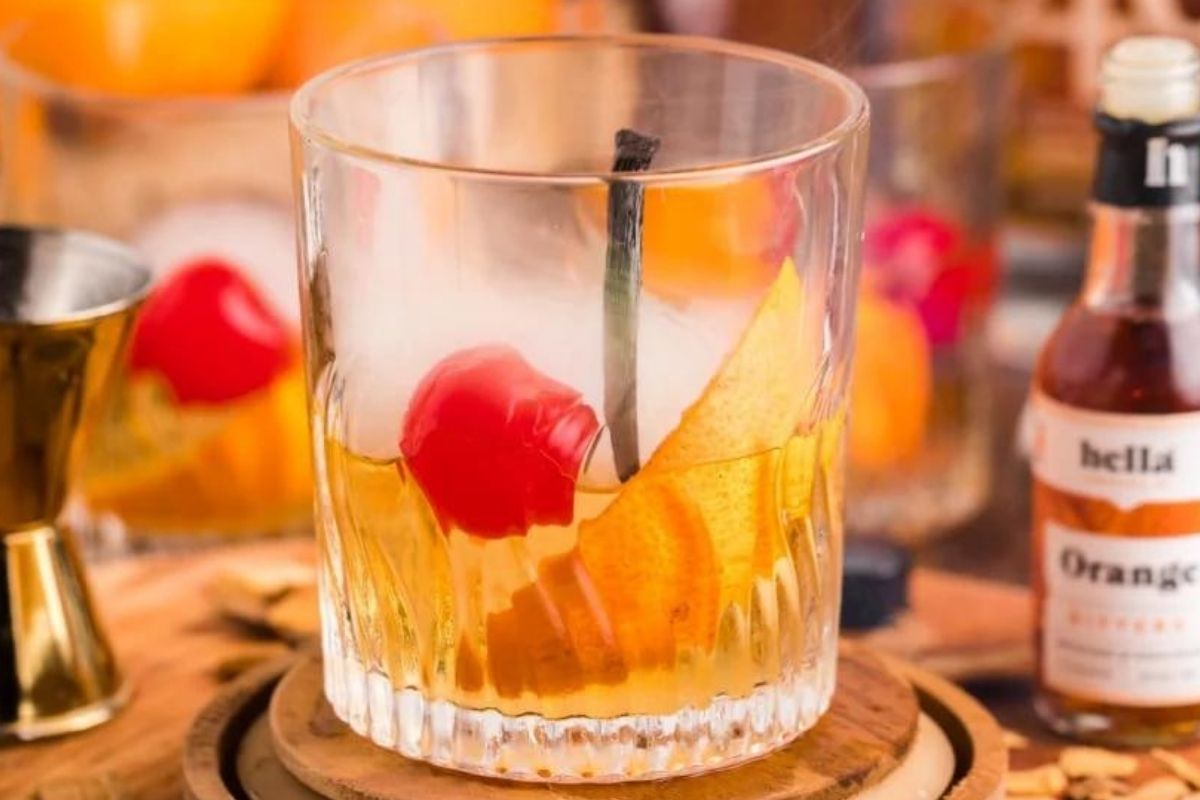 A glass of whiskey with ice, garnished with an orange peel and a cherry, surrounded by smoked cocktail-making ingredients.