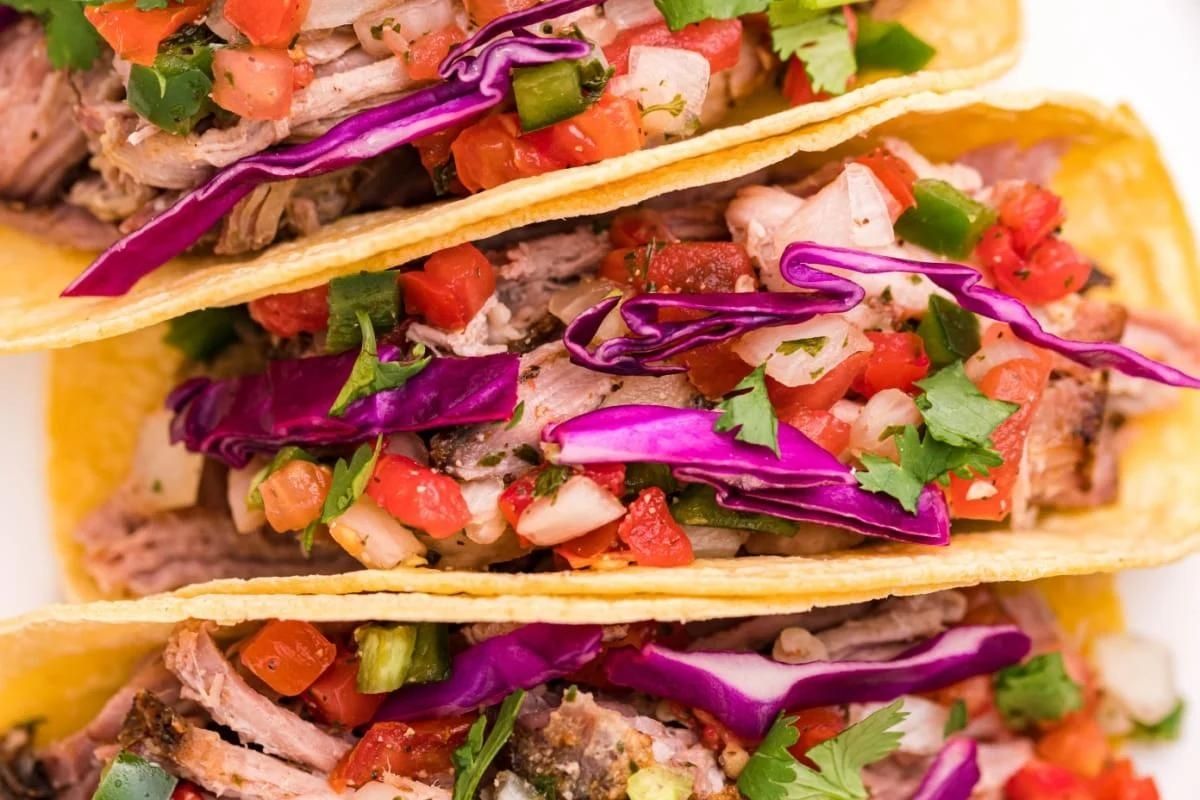 Three tacos filled with smoked pulled pork, red cabbage, diced tomatoes, and chopped cilantro served in yellow corn tortillas.