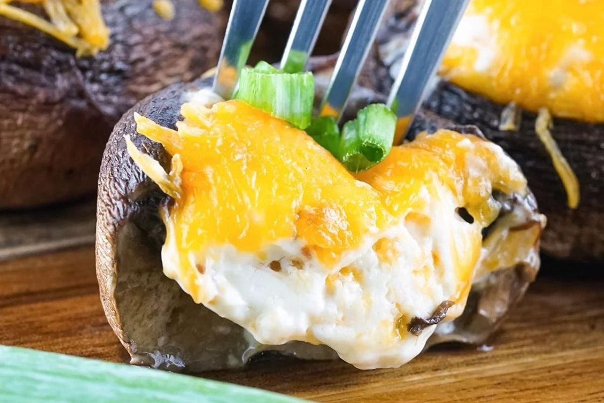 A fork piercing a cheesy stuffed mushroom topped with melted smoked cheddar cheese and green onions on a wooden surface.