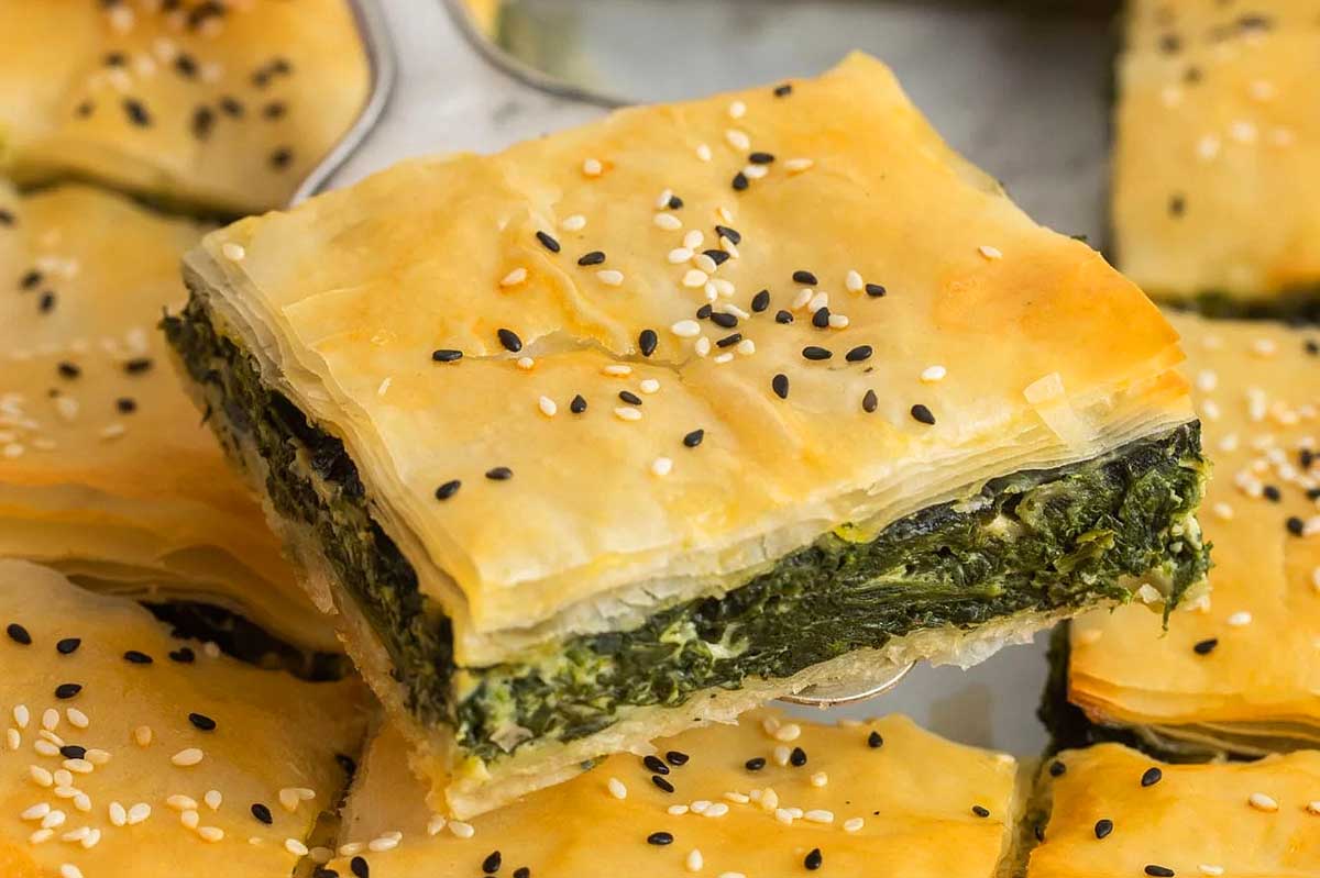 A piece of spinach and cheese pie with flaky golden pastry topped with sesame seeds, prepared using sheet pan recipes, with more pieces in the background.