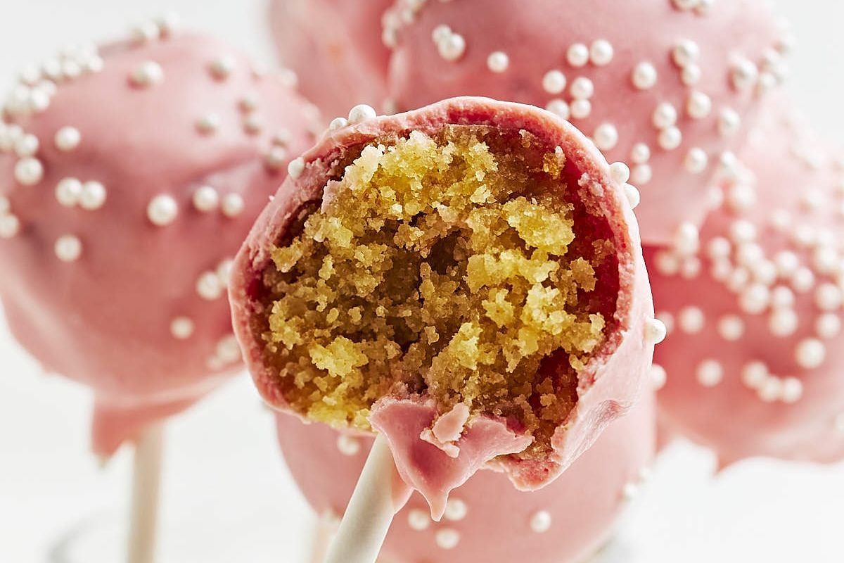 Close-up of cake pops with pink icing and white sprinkles, looking just like those from copycat recipes. One cake pop is partially eaten, revealing yellow cake inside.