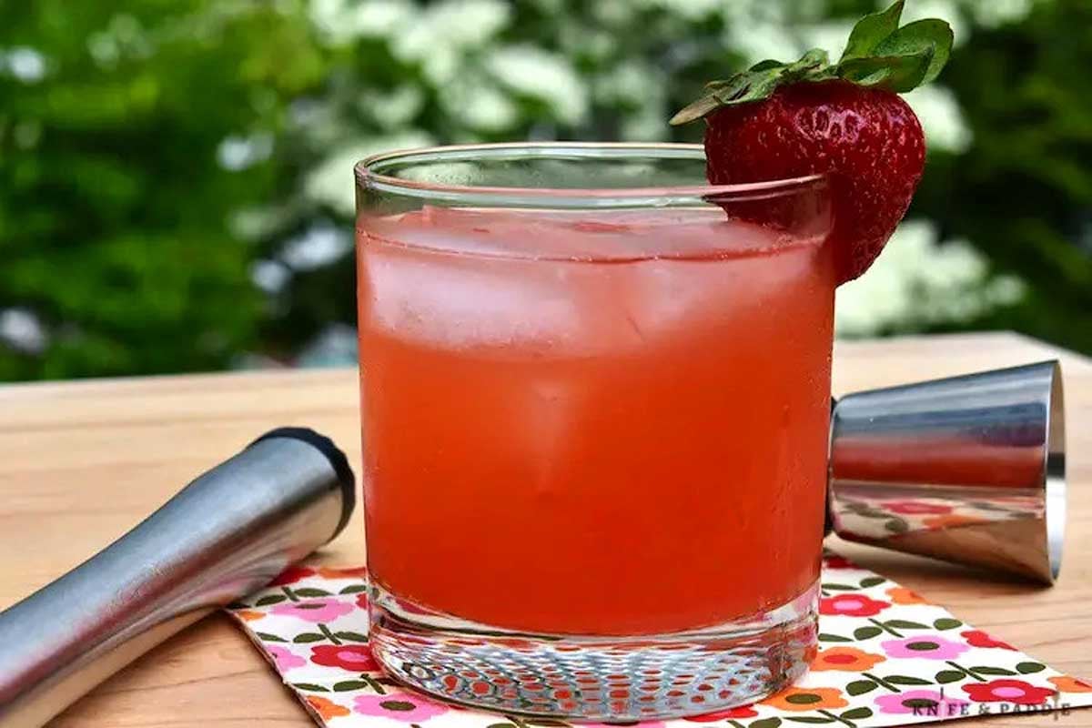 A glass of strawberry-rhubarb cocktail garnished with a whole strawberry, with a shaker and cocktail stirrer beside it on a wooden table.