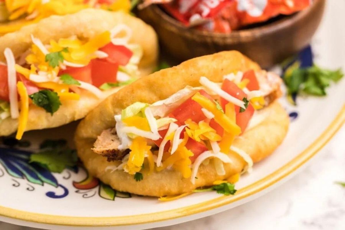 Two chicken dinner tacos with shredded cheese, lettuce, and diced tomatoes on a decorative plate.