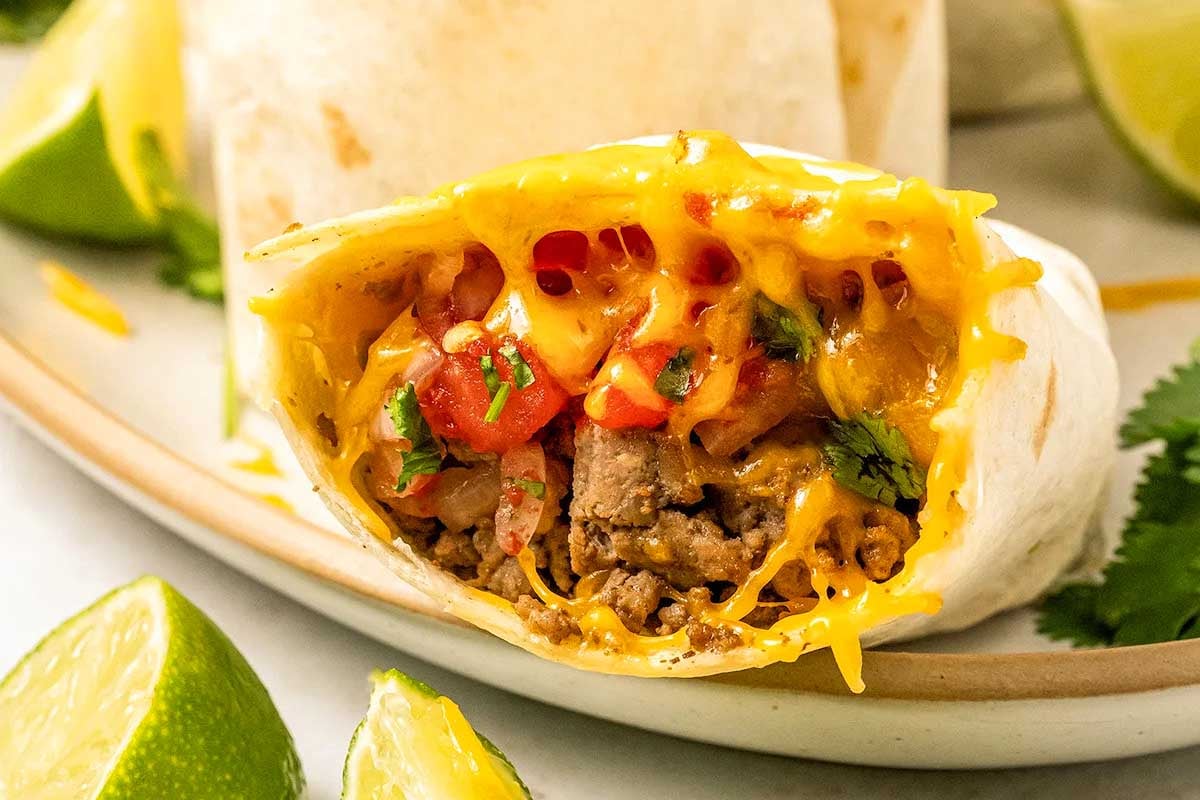 A close-up image of a cut burrito filled with beef, cheese, and tomatoes, surrounded by lime wedges and cilantro on a plate, inspired by Taco Bell copycat recipes.