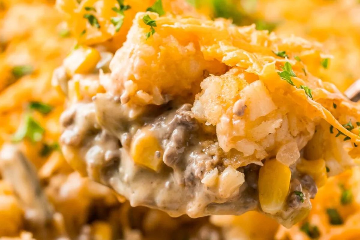 A close-up of a creamy casserole with a spoon lifting a portion, showing layers of ground beef, corn, and melted cheese mixed with can of cream soup, garnished with chopped parsley.