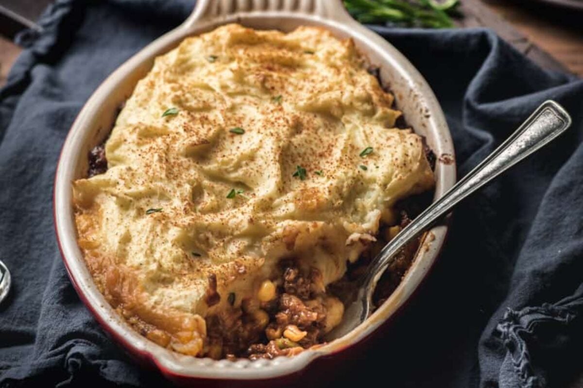 Shepherd’s pie in a baking dish with a spoon, topped with golden mashed potatoes and herbs, made easy by using frozen vegetables for a quick and hearty meal.