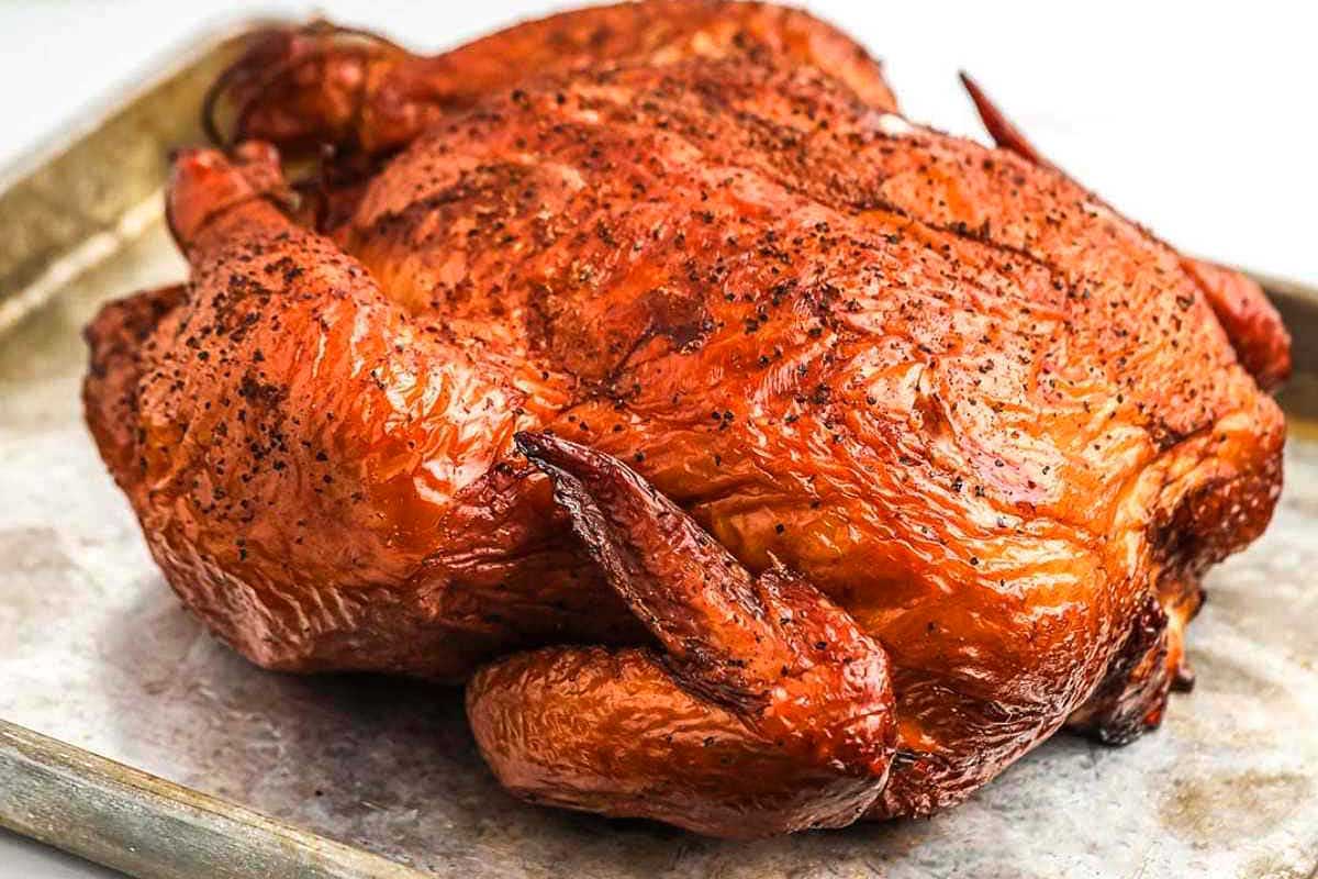 A smoked chicken with a crispy, golden-brown skin, seasoned with herbs, displayed on a metal tray.
