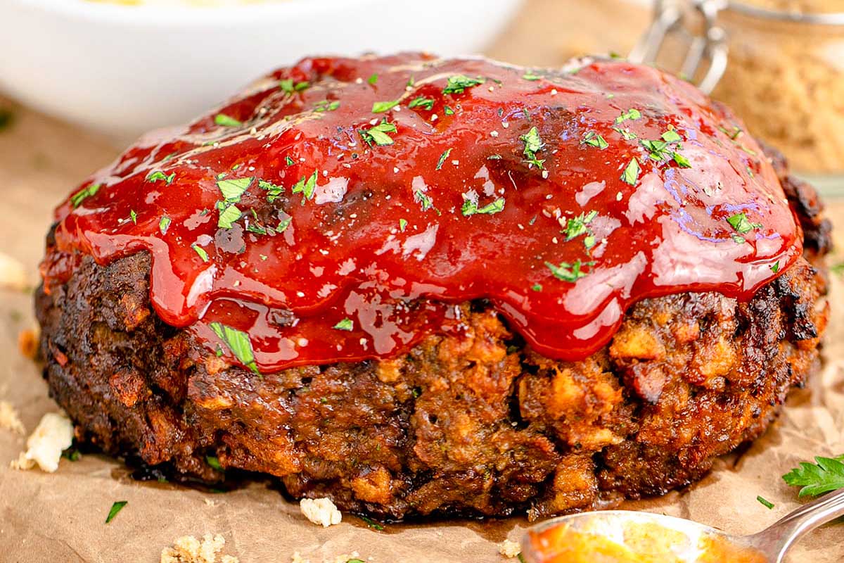 A close-up image of a ketchup-glazed meatloaf topped with fresh herbs on a wooden table.