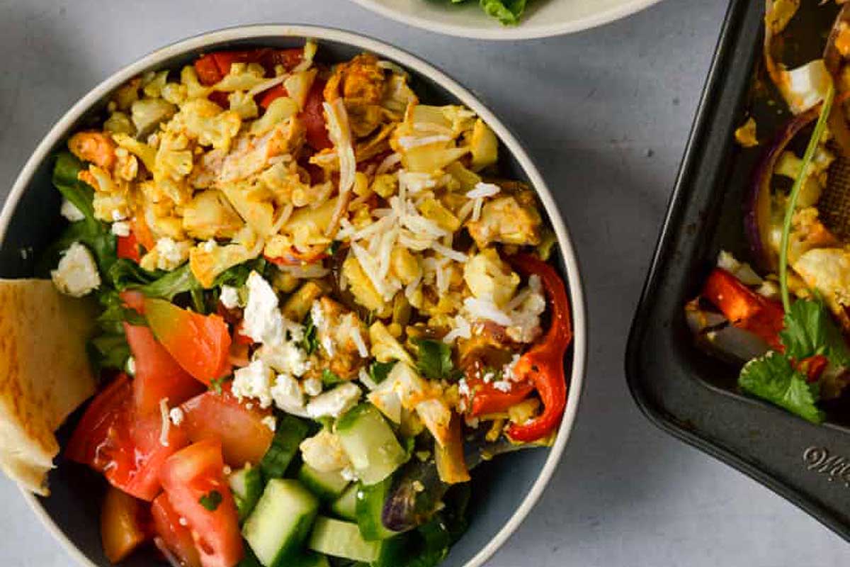 A bowl of Greek salad with tomatoes, cucumbers, peppers, feta cheese, and chicken, next to a sheet pan of similar ingredients.