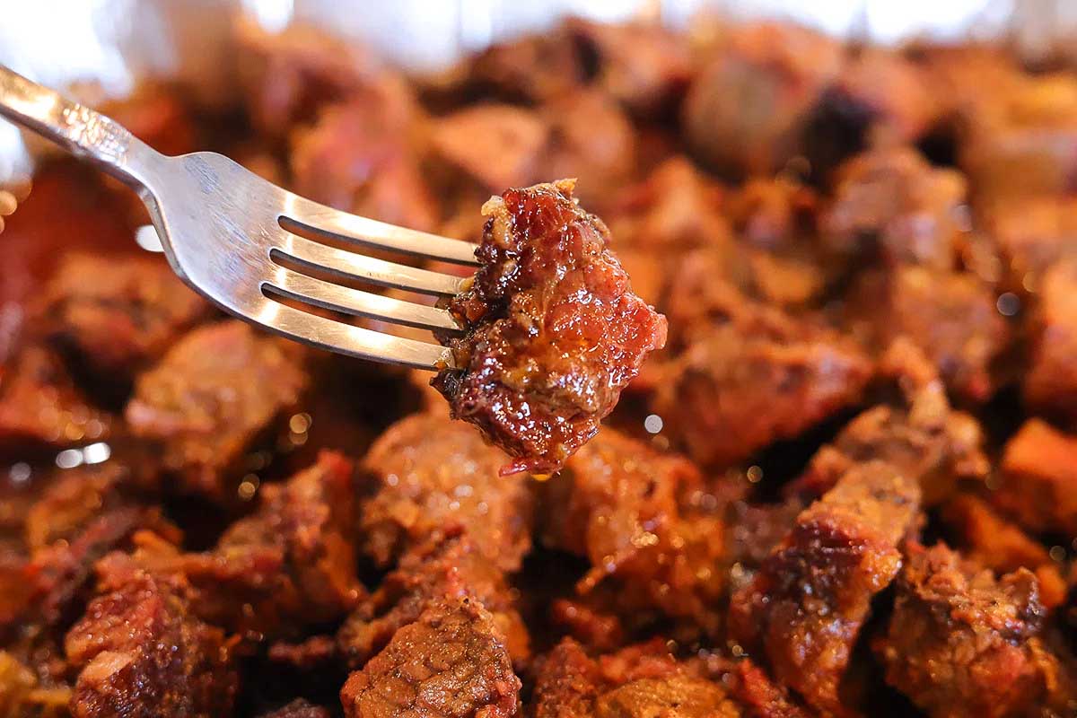 A close-up image of a fork spearing a juicy, caramelized piece of meat from smoker recipes, surrounded by several other cooked meat pieces.