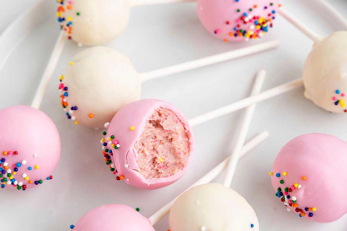 A plate of pink and white cake pops with colorful sprinkles, one partially bitten, inspired by recipes that use circus animal cookies, on a white background.