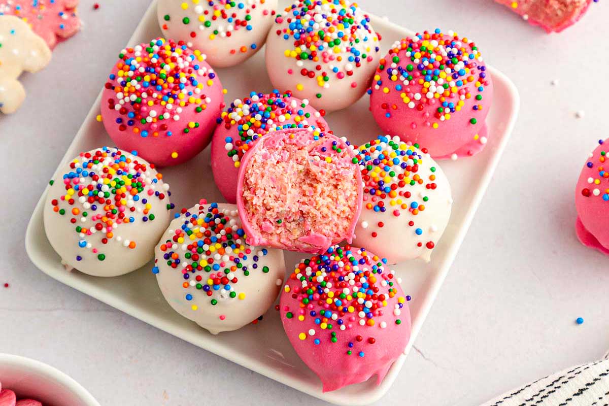 A plate of pink and white cake balls covered with colorful sprinkles and circus animal cookies, one cut in half showing a pink interior.