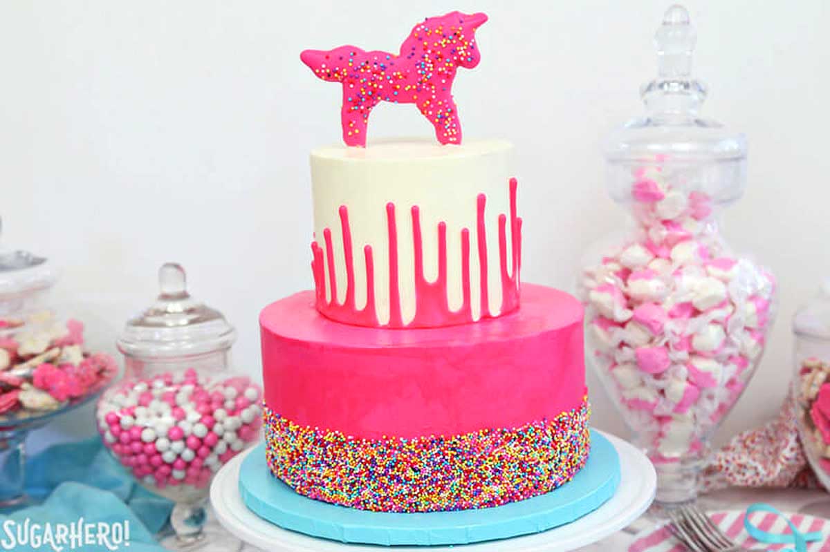 A two-tiered celebration cake with pink frosting and multicolored sprinkles, topped with a pink fondant unicorn and circus animal cookies, displayed on a table with candy jars.