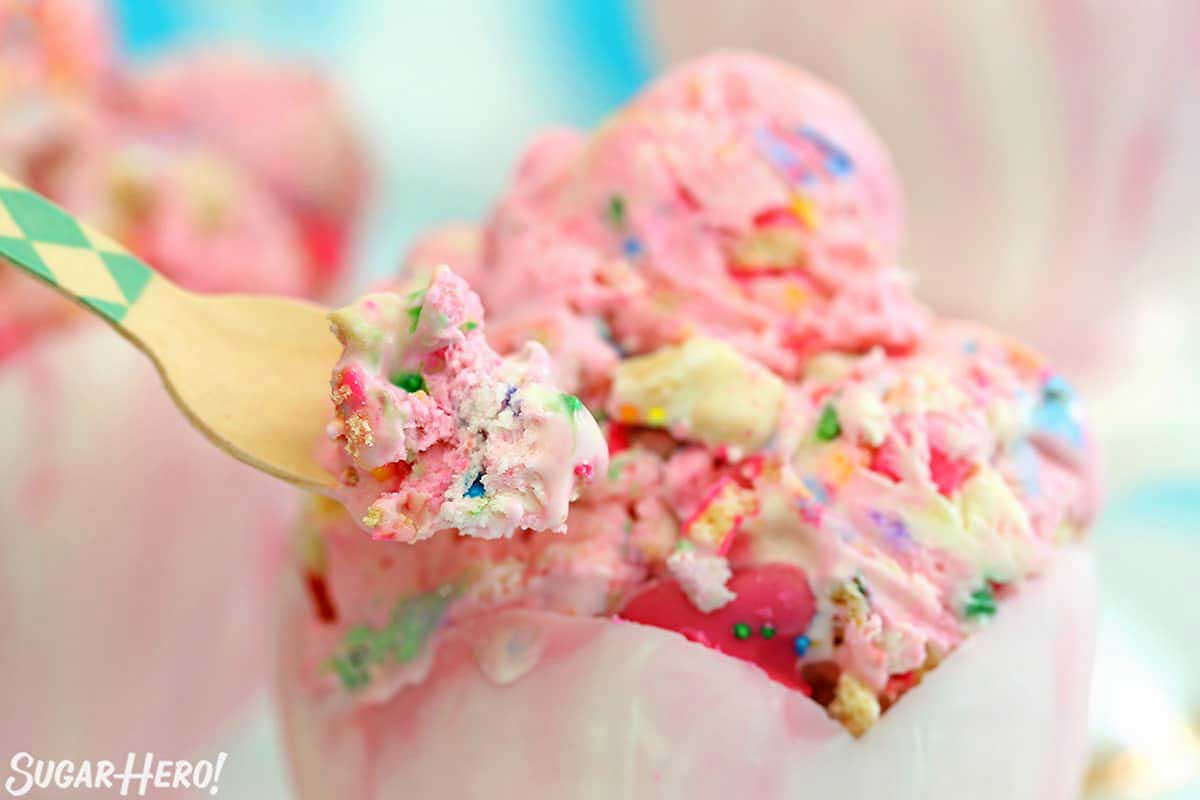 A spoon scooping colorful birthday cake ice cream topped with circus animal cookies and sprinkles from a cup.