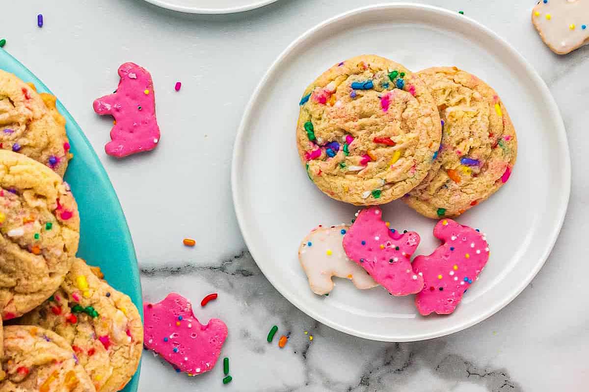 A marble countertop with plates of colorful sprinkle cookies and pink frosted circus animal cookies.