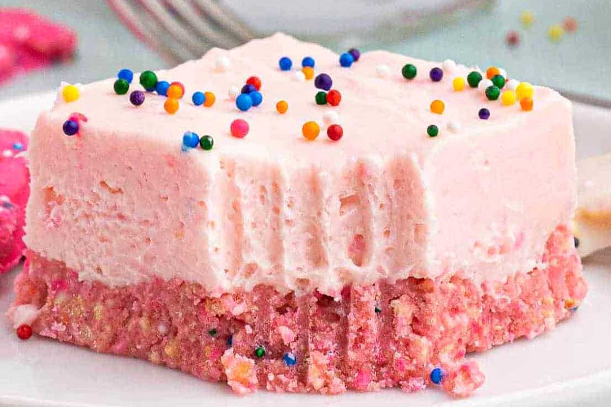 A slice of pink strawberry cake with colorful Circus Animal Cookie sprinkles on top, served on a white plate with more cake in the background.