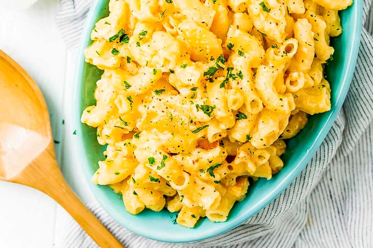 A bowl of creamy macaroni and cheese from slow cooker recipes, garnished with herbs, served on a light blue dish with a wooden spoon beside it.