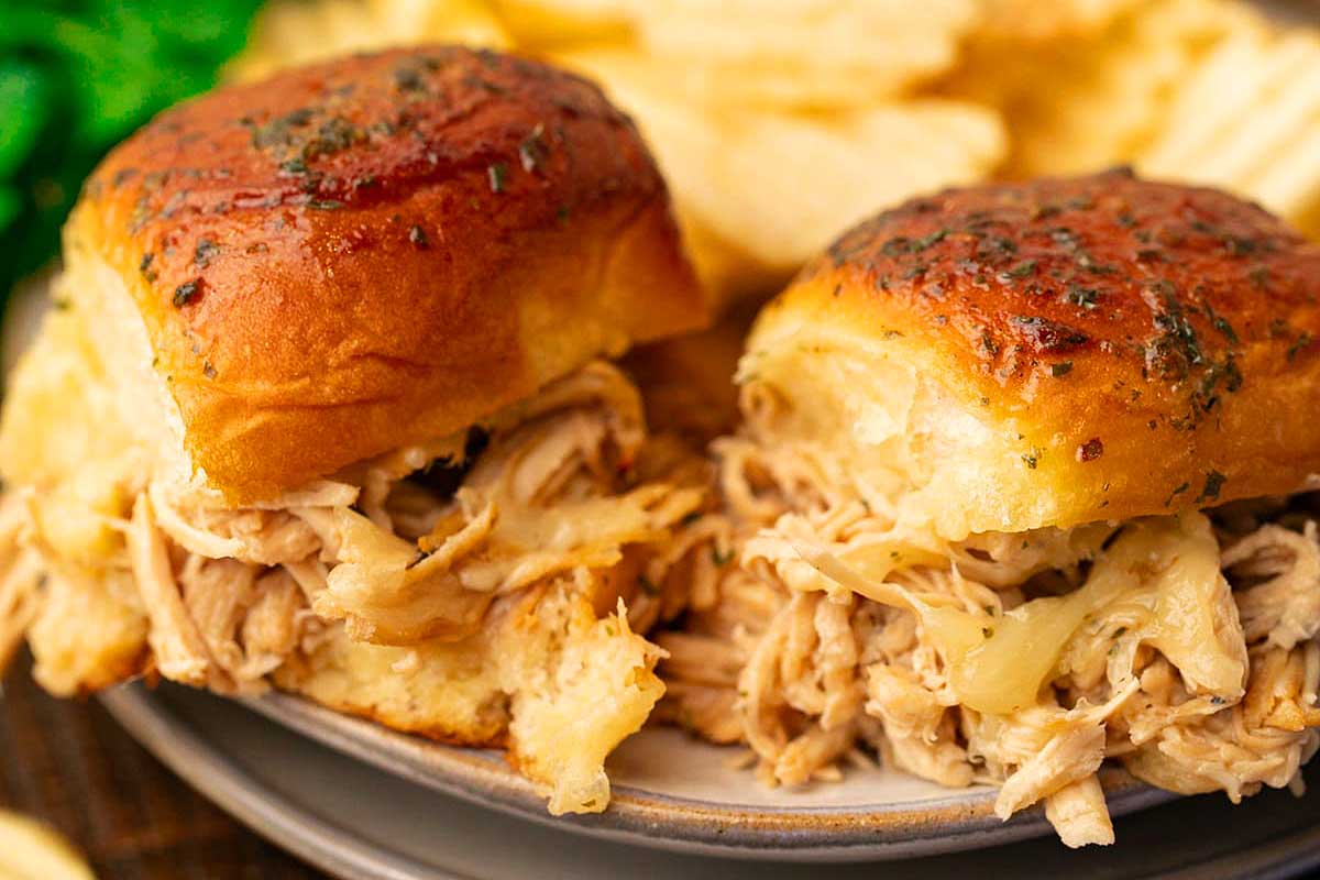Close-up of slow cooker pulled pork sandwiches on a plate with potato chips. Sandwich buns are topped with herbs.