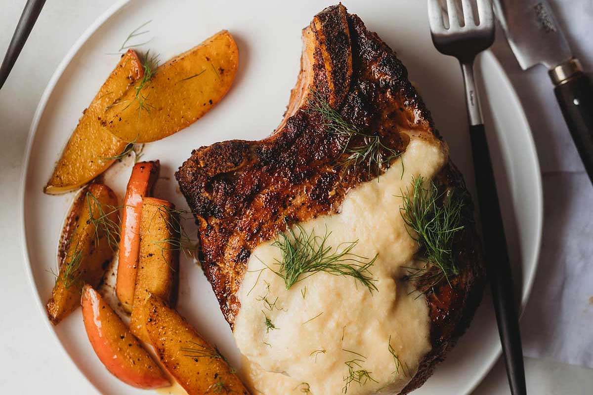 Smoked pork chop topped with sauce and garnished with dill, served with roasted carrots and peaches on a white plate.