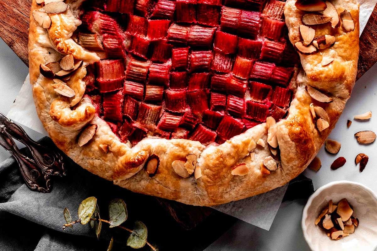 A freshly baked rhubarb pie with a lattice top, surrounded by almonds and a small bowl of whipped cream, perfect for those exploring new rhubarb recipes.