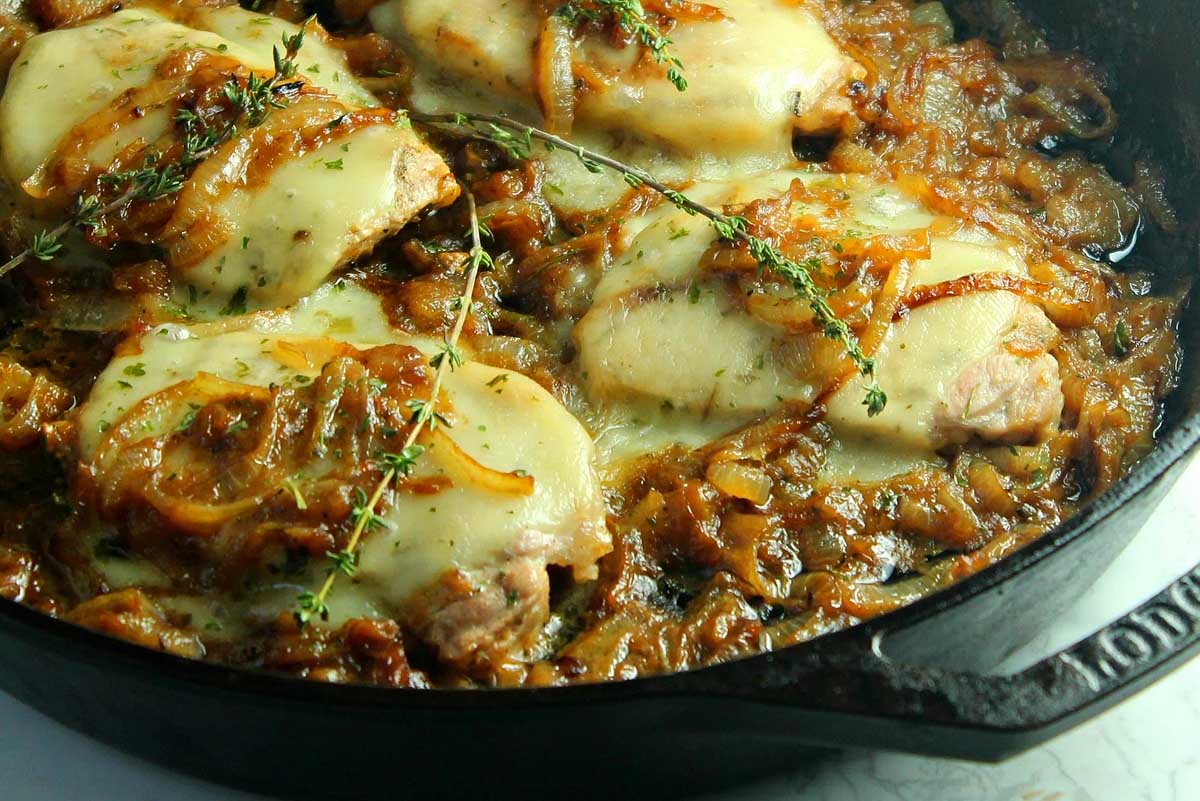 Pork chop recipes: Chicken thighs topped with melted cheese in a skillet, garnished with thyme and surrounded by a rich, onion-laden sauce.