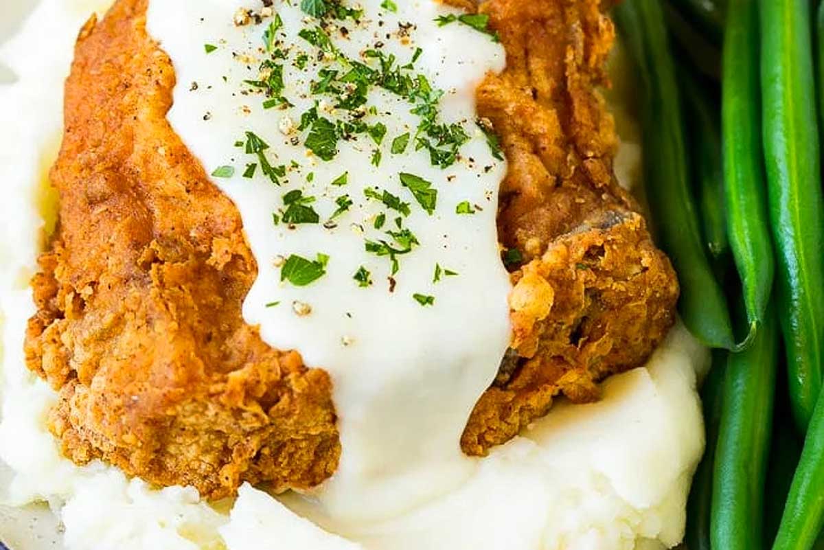 A plate of fried pork chop with creamy gravy on top, served with mashed potatoes and green beans, garnished with parsley.