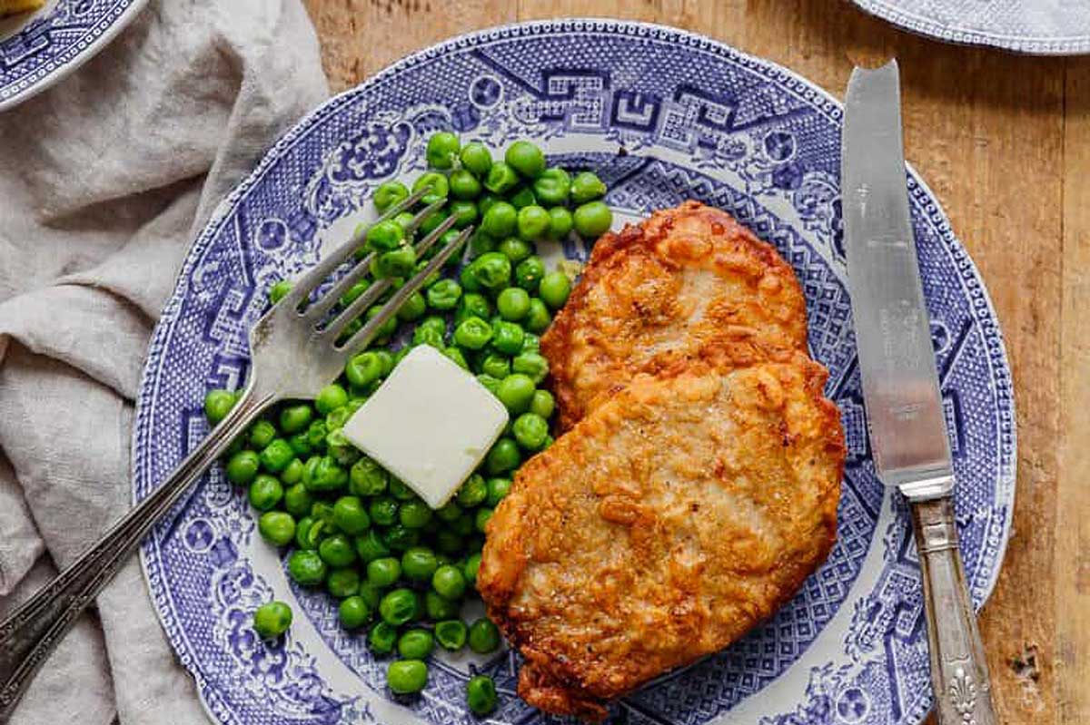 A plate with two fried potato pancakes, a pork chop, and green peas, accompanied by a fork and knife, on a blue and white patterned plate.