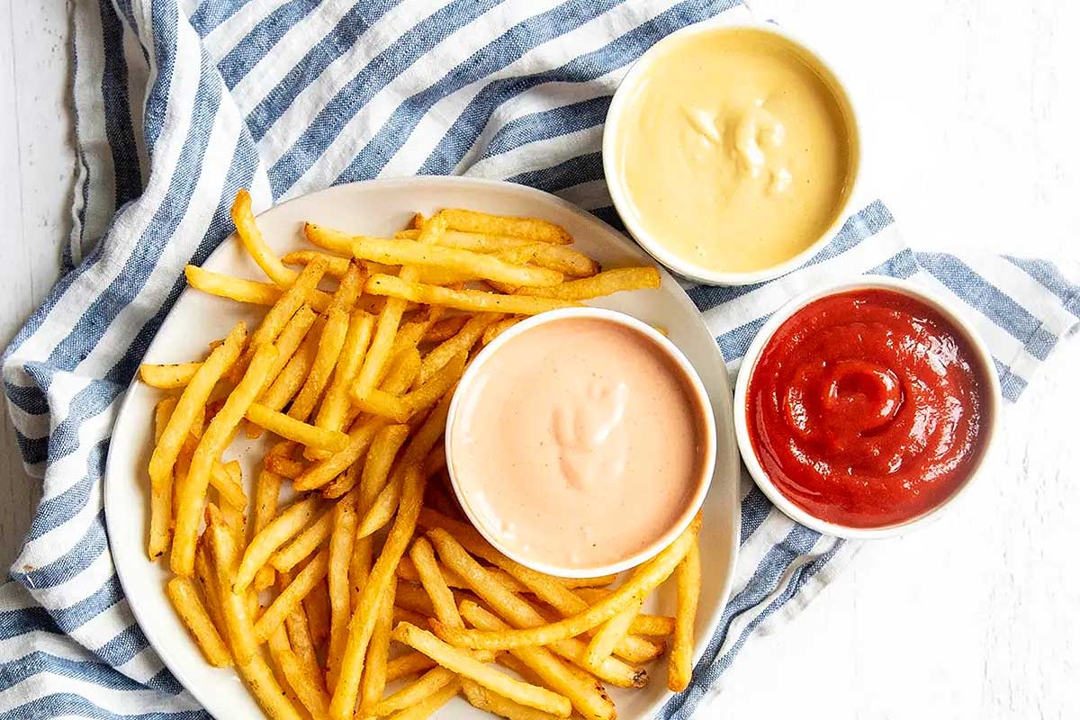 Plate of crispy french fries with three dipping sauces: ketchup recipes, mayonnaise, and a pink sauce, arranged on a striped napkin.