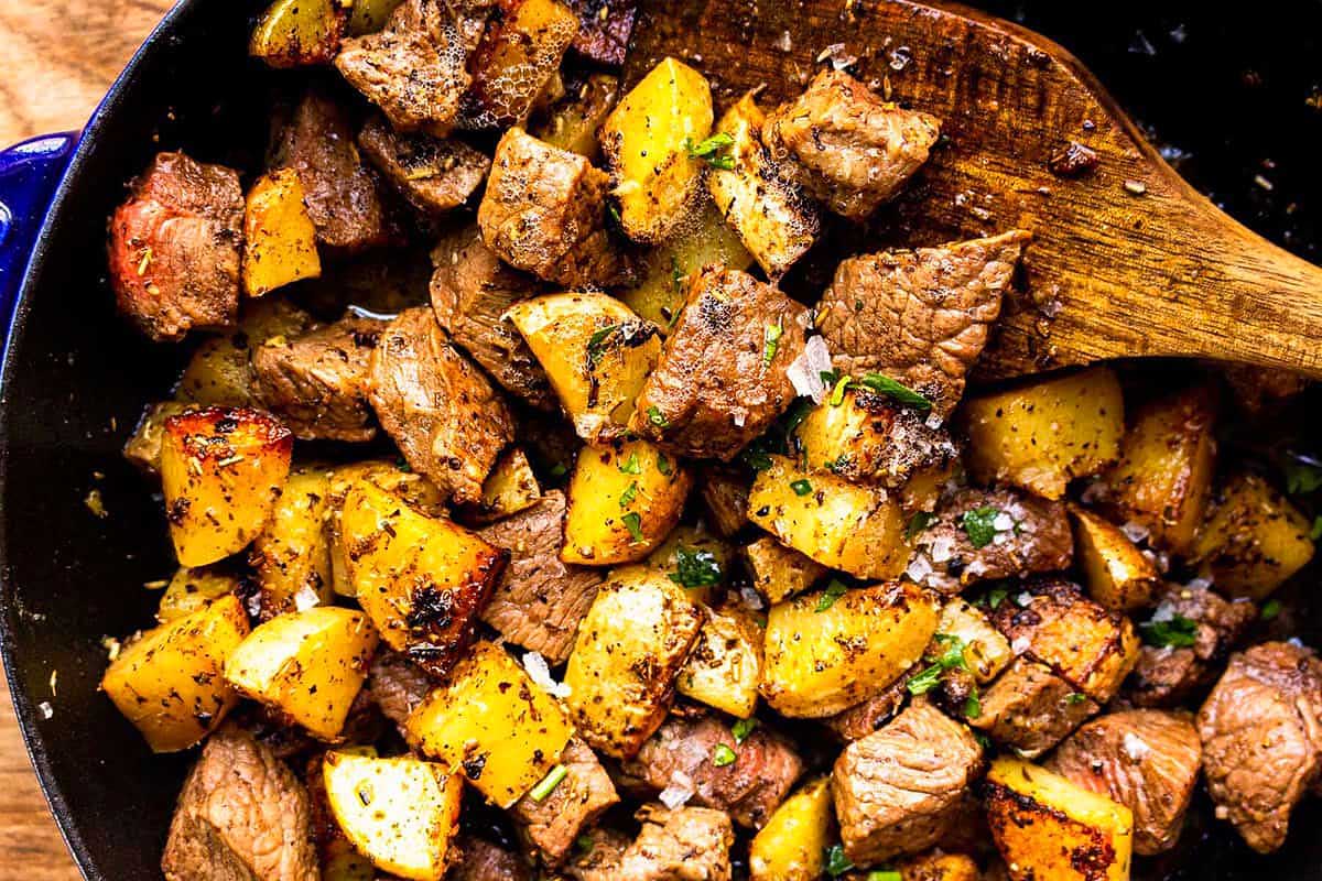 Cubes of seasoned steak and potatoes are stirred in a skillet with a wooden spoon, creating a hearty meal ready in 30 minutes or less.