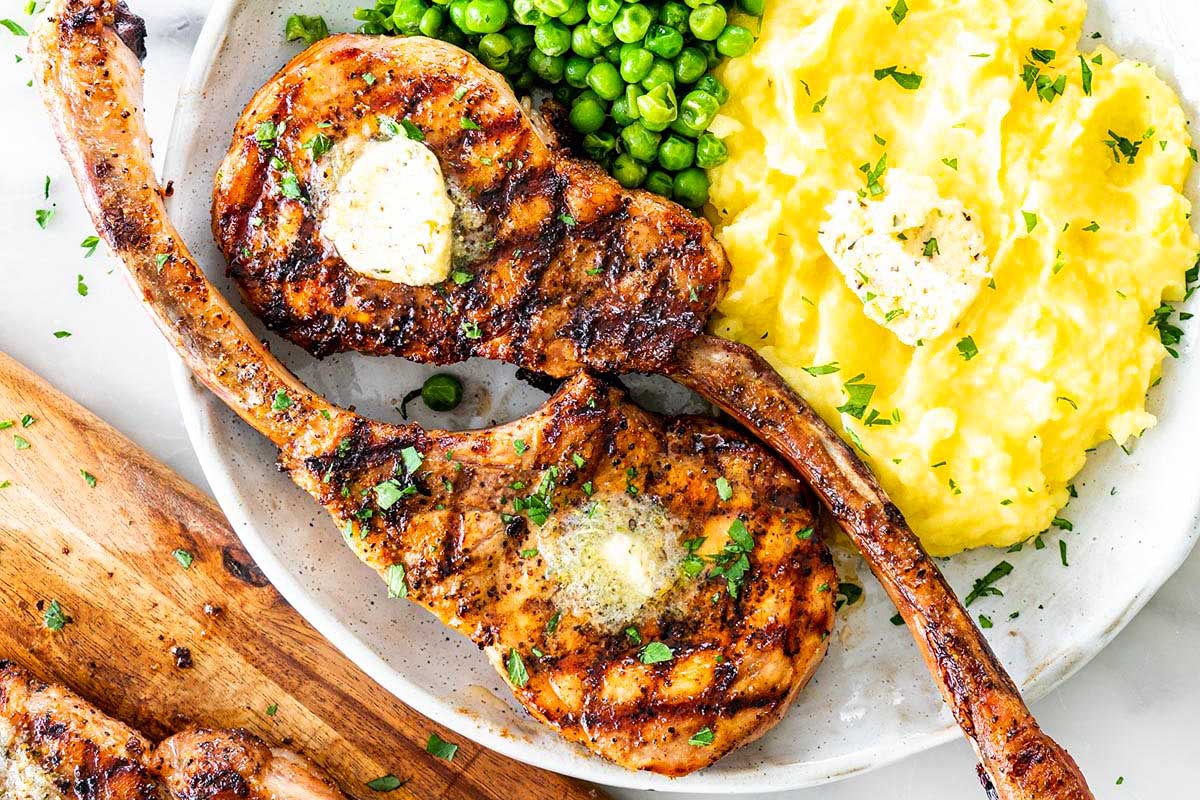 Grilled pork chop recipes with herb butter on top, served with mashed potatoes and peas on a white plate.
