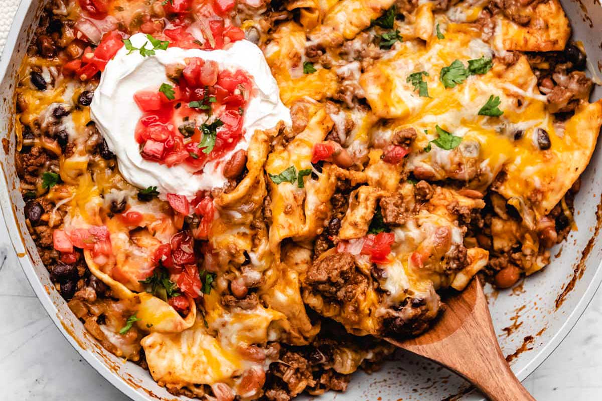 A skillet filled with beef and bean nachos topped with melted cheese, tomatoes, and a dollop of sour cream, prepared in 30 minutes or less, with a wooden spoon.