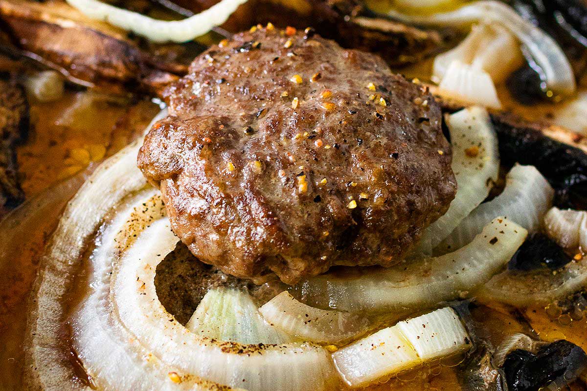 Juicy grilled hamburger patty on a sheet pan with sliced onions and mushrooms, all featuring visible seasoning.