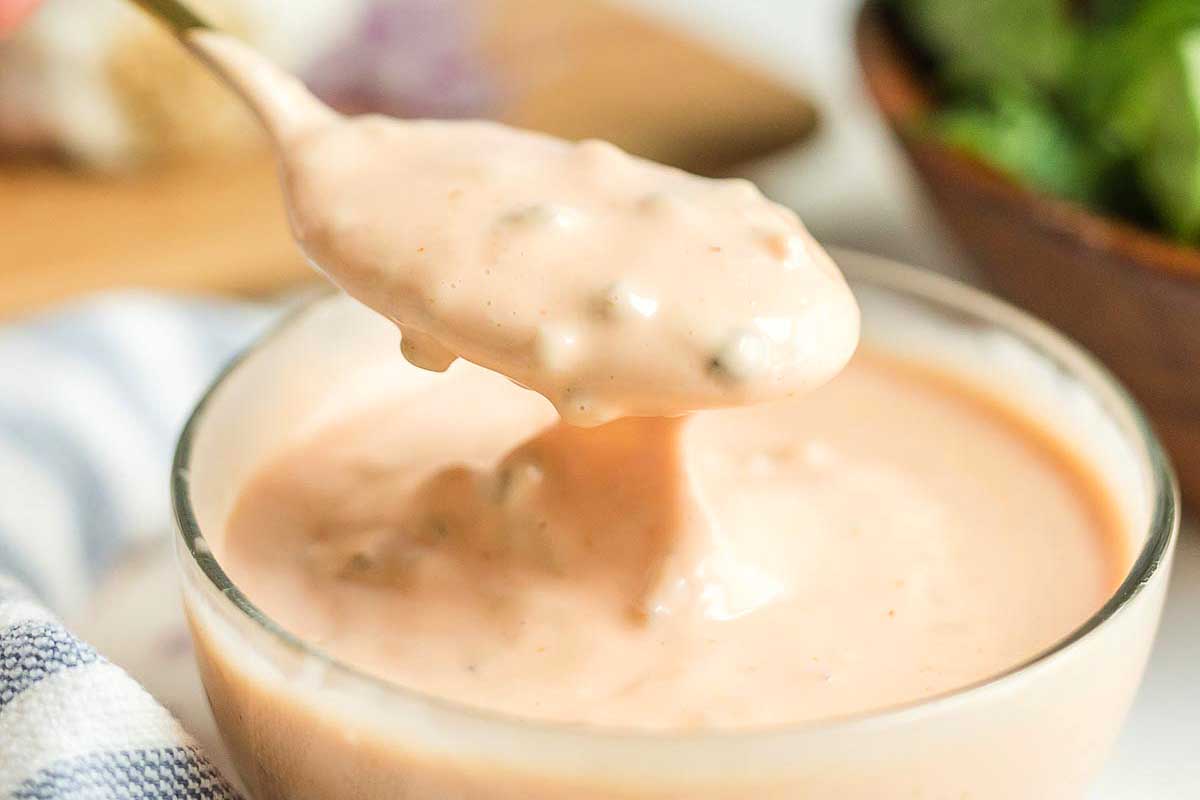 A close-up image of creamy ketchup dressing being poured from a spoon into a glass bowl with a backdrop of fresh greens.