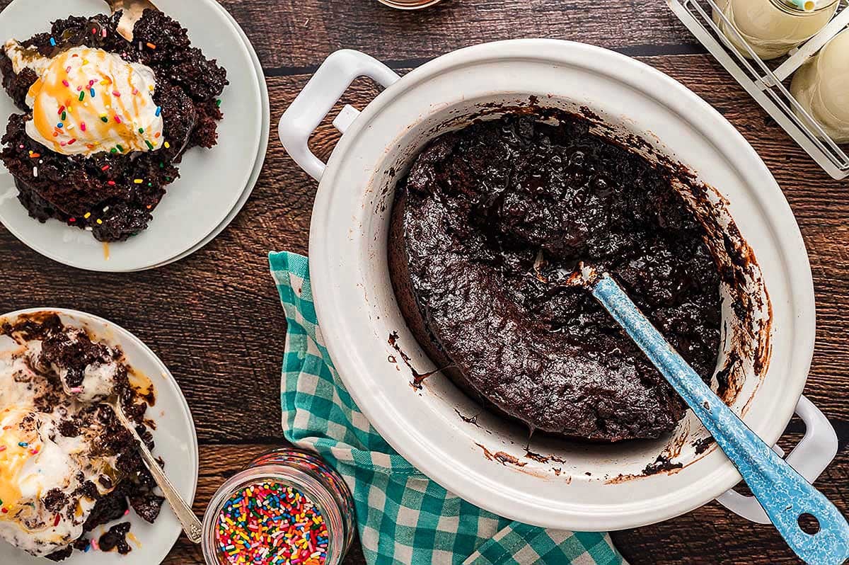 A homemade chocolate cake made with pudding mix in a white baking dish with pieces served alongside ice cream and sprinkles.