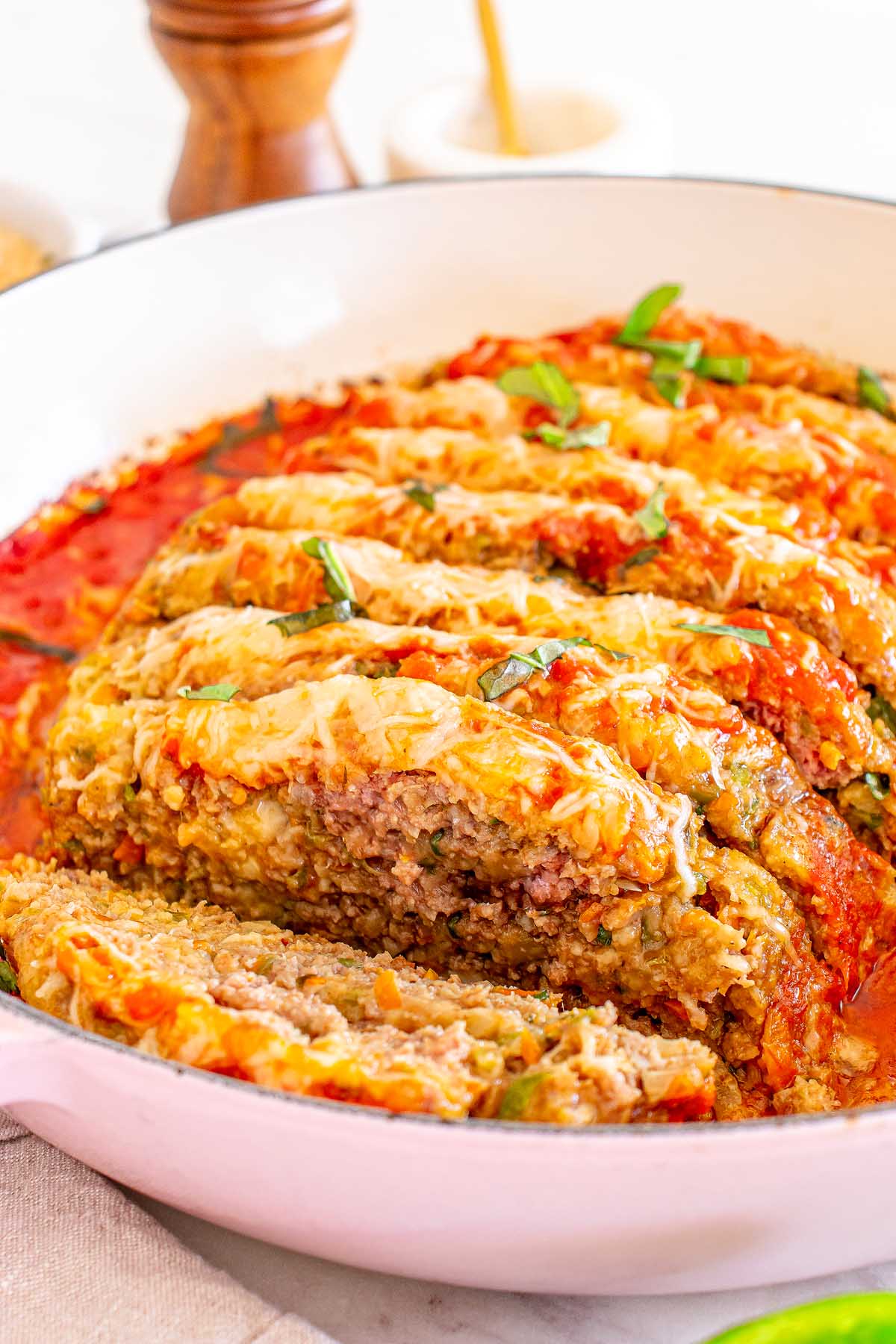 A sliced Italian meatloaf topped with tomato sauce and melted cheese, garnished with chopped green herbs in a white dish.