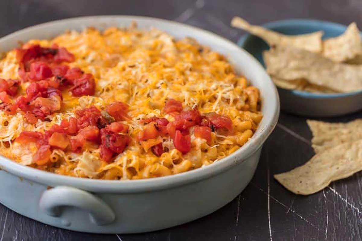 A baked Velveeta cheese dish topped with diced tomatoes in a ceramic bowl, served with tortilla chips on a dark surface.