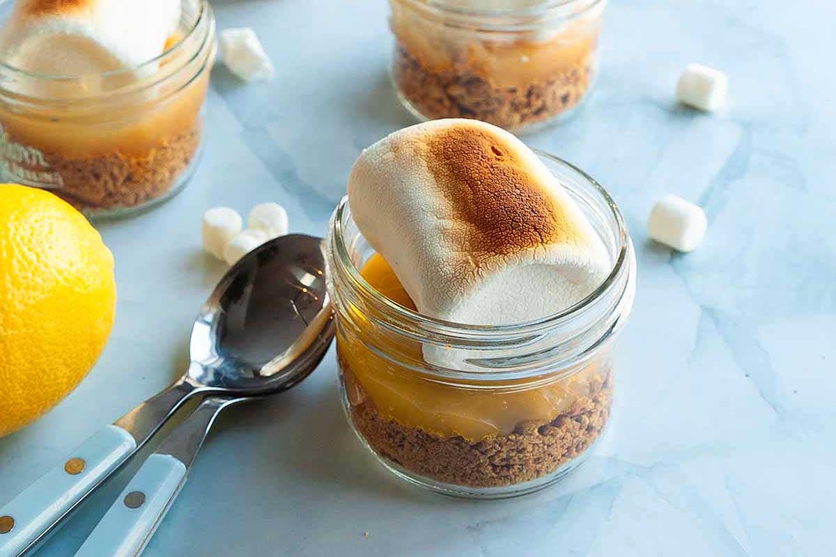 A lemon curd meringue dessert in a glass jar with a toasted marshmallow on top, accompanied by a spoon, some marshmallows, and a lemon on a marble surface.