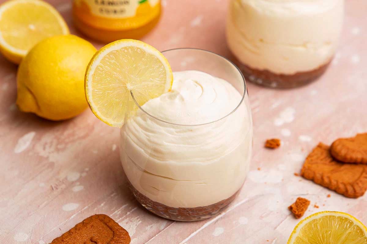 A glass of lemon curd mousse topped with whipped cream and a lemon slice, surrounded by whole lemons and cookies on a pink surface.