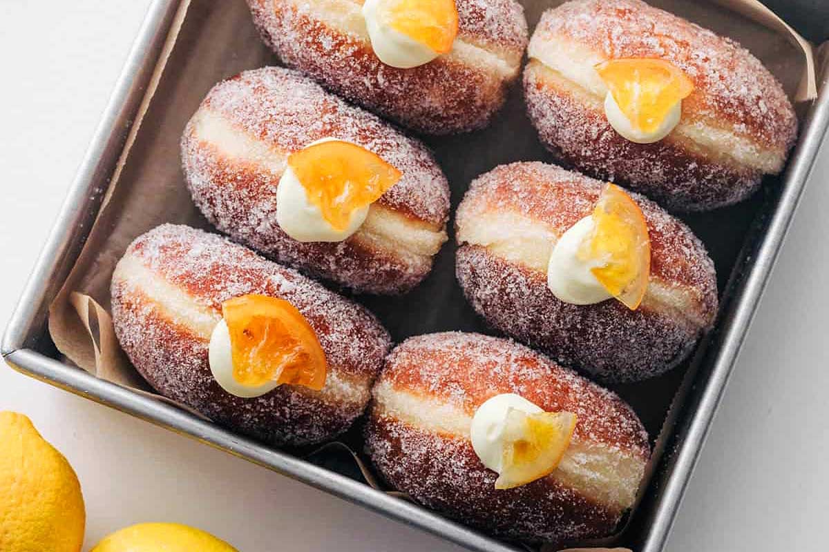 Sugar-dusted doughnuts, some topped with lemon curd and candied lemon, arranged neatly in a metal tray on a white surface.