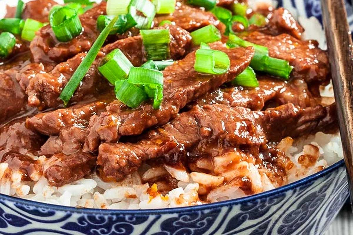 Close-up of beef teriyaki over rice garnished with chopped green onions in a blue patterned bowl, ready in 30 minutes or less.