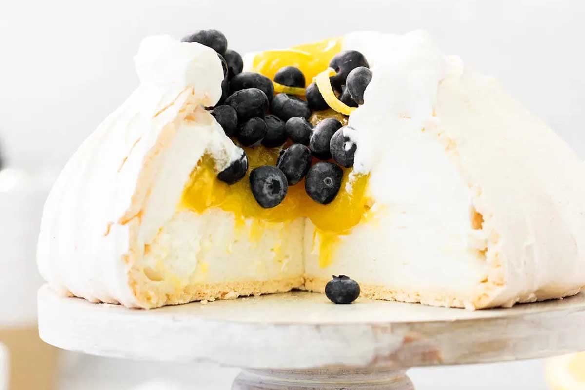A pavlova topped with lemon curd-recipes and fresh blueberries on a white cake stand.