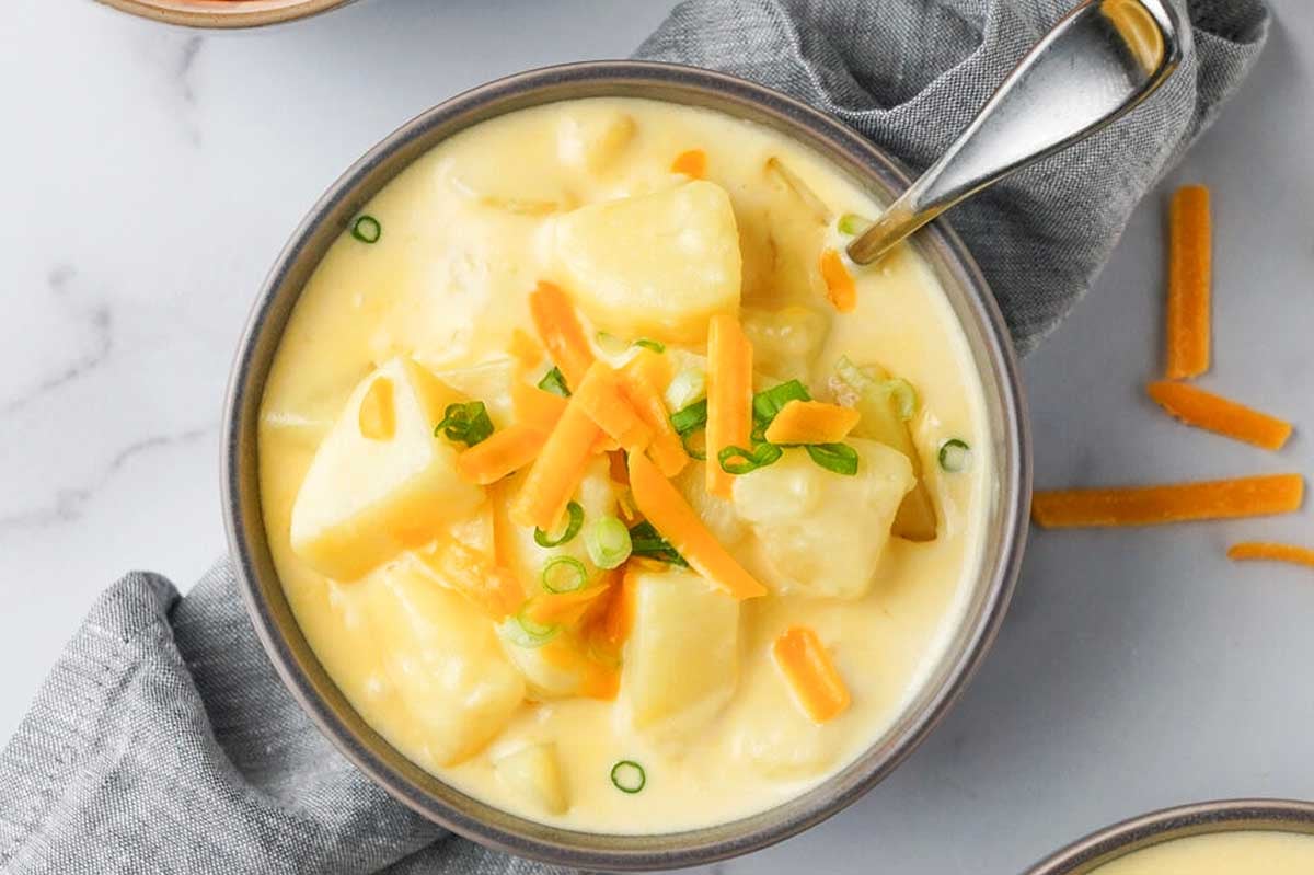 A bowl of creamy potato soup garnished with shredded Velveeta cheese and chopped green onions, served with a spoon on a marble surface.