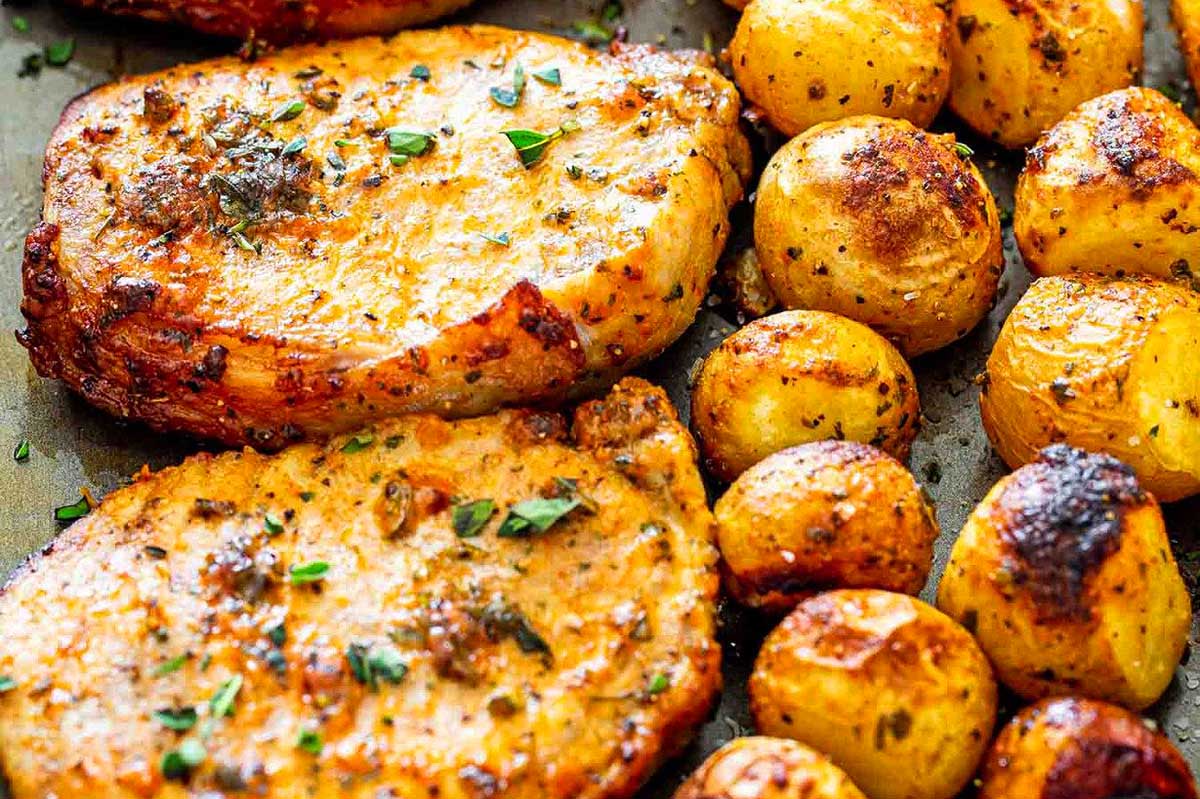 Grilled pork chops garnished with herbs alongside roasted golden potatoes on a baking sheet, all ready in 30 minutes or less.