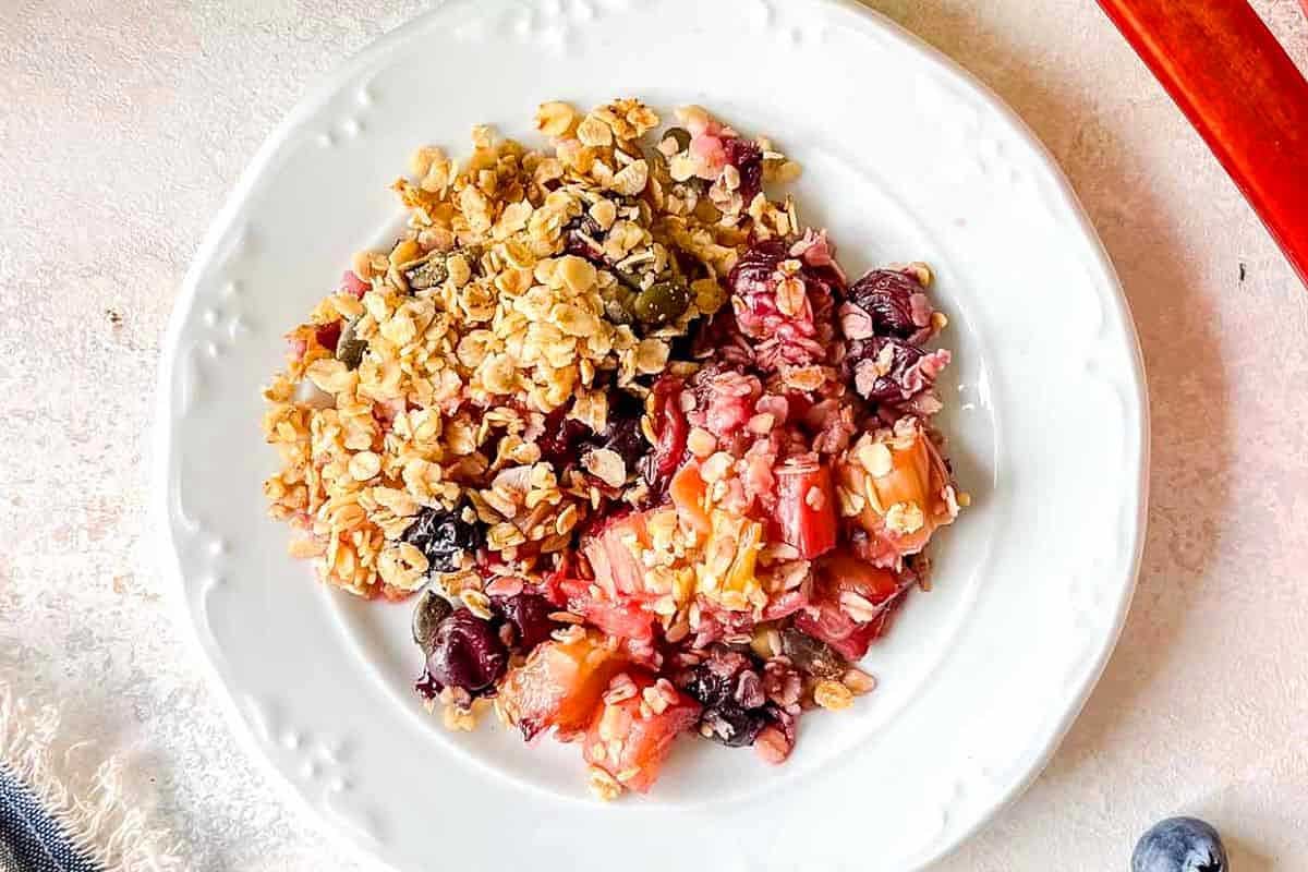 A plate of fruit crumble with mixed berries, chopped apples, and rhubarb, topped with a golden-brown oat crumble, served on a white plate with a red spoon on the side