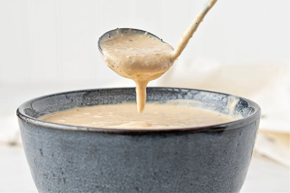 A spoon drizzling creamy sauce into a gray ceramic bowl filled with the same sauce—perfect for elevating your favorite ketchup recipes.