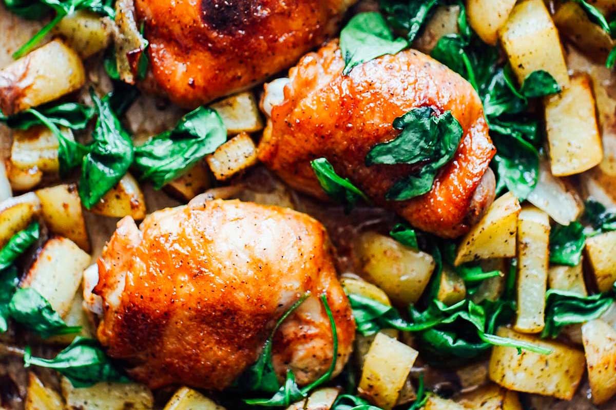 Roasted chicken thighs with golden potatoes and fresh spinach leaves in a sheet pan.
