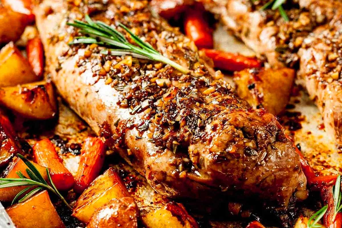 Roasted pork tenderloin garnished with rosemary and surrounded by caramelized apples and vegetables on a sheet pan.