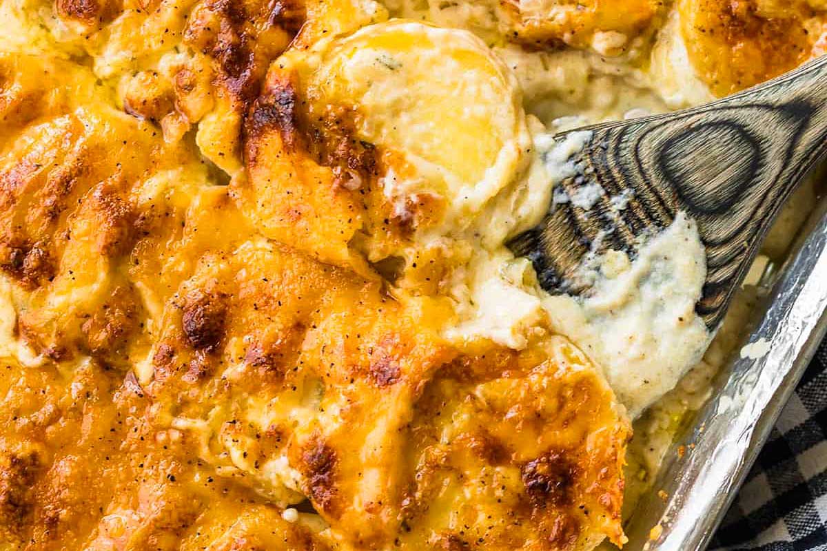 A close-up of a cheesy scalloped potatoes dish in a sheet pan, showing layers of sliced potatoes covered in melted cheese and a serving spoon.