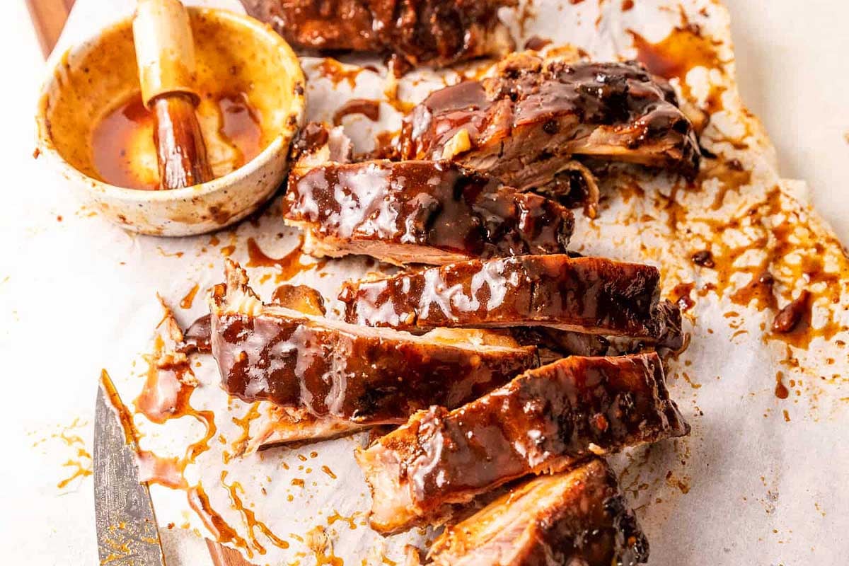Sliced barbecue ribs cooked using slow cooker recipes, covered in sauce with a basting brush and sauce bowl on a parchment-lined surface.