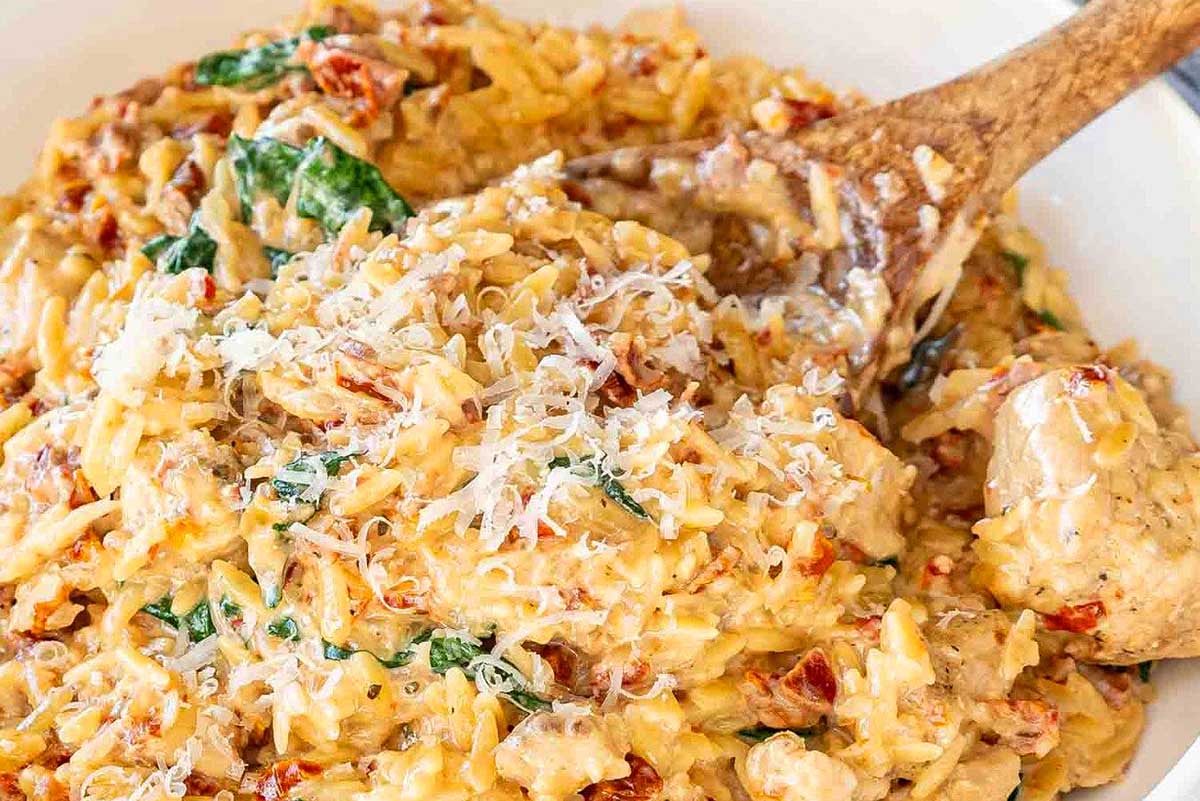 A creamy risotto with spinach, sun-dried tomatoes, and chicken, garnished with grated parmesan cheese, prepared in a slow cooker and served in a white bowl with a wooden spoon.